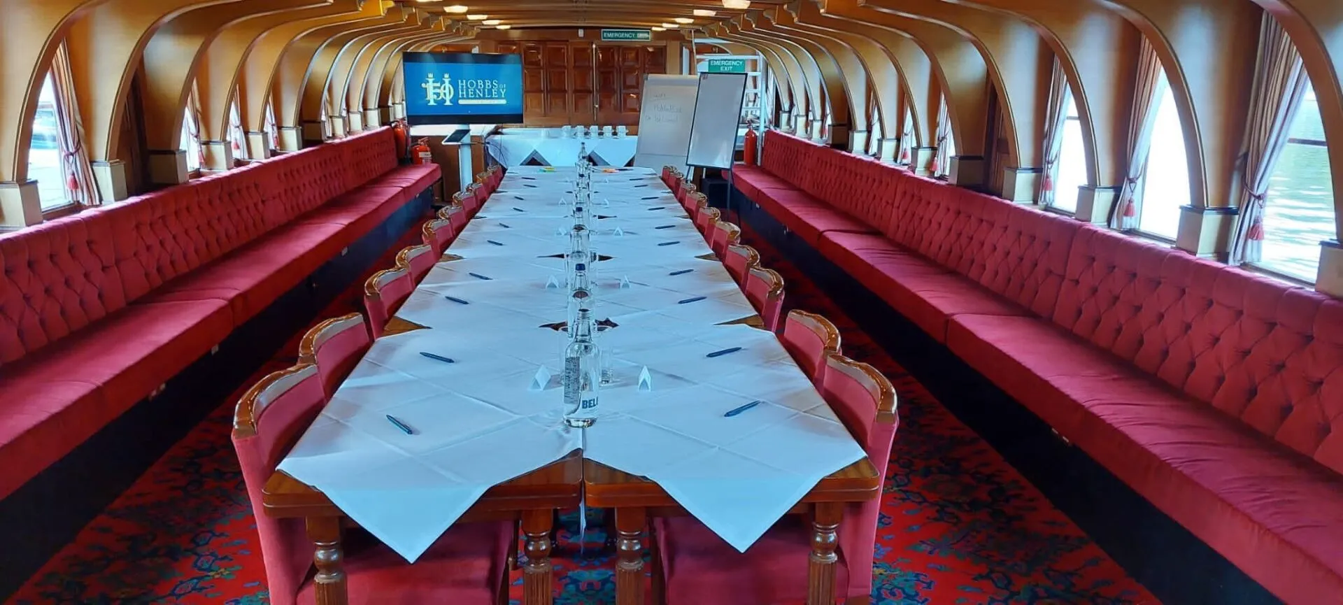 Inside one of the Hobbs of Henley passenger boats, set up for a corporate event. The table is laid for 20 people with bottled water and pens at the ready. There is a TV and whiteboards set up at the back of the boat.