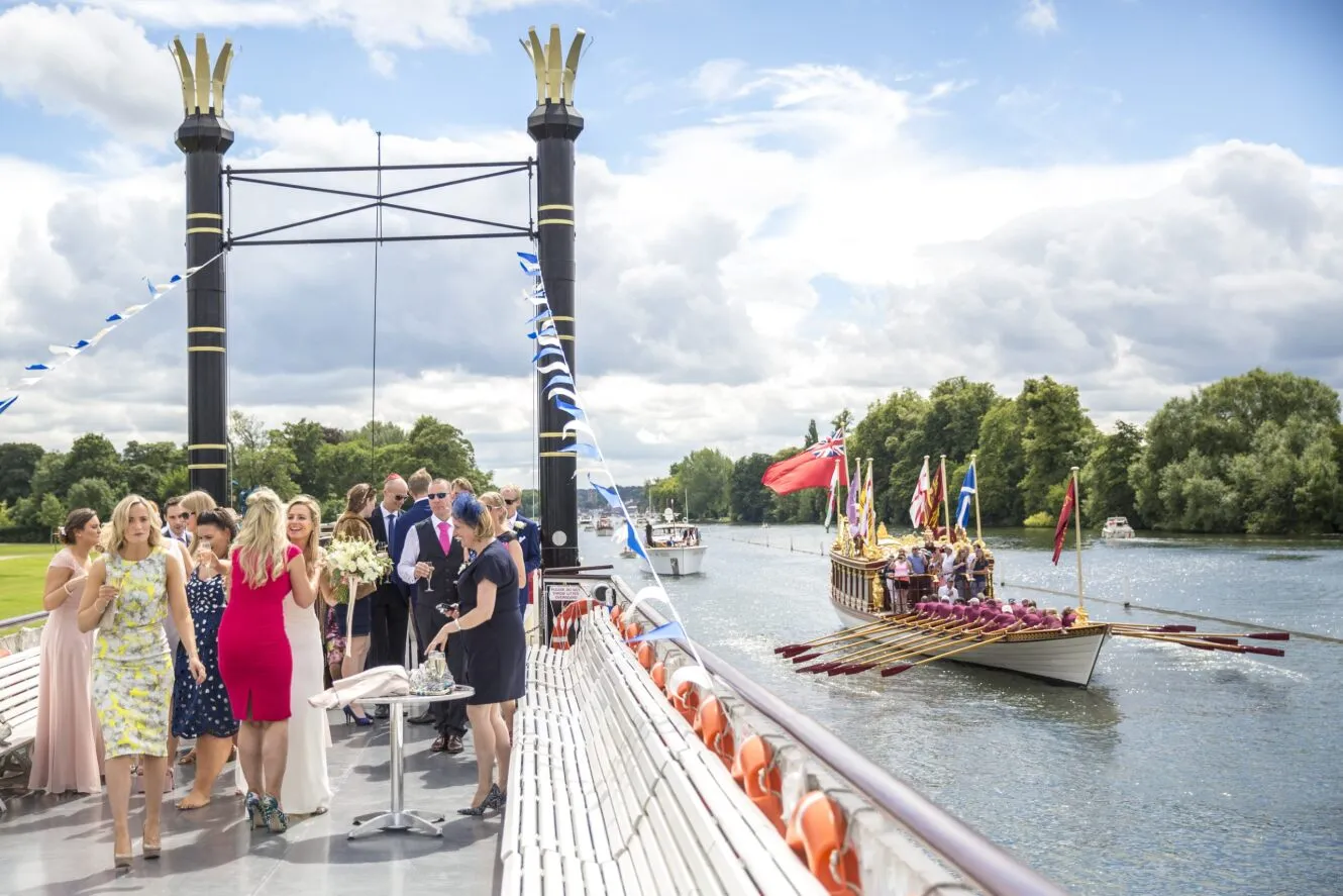 A wedding celebration aboard The New Orleans boat. Passengers are enjoying the outside of the boat, while cruising along the river thames with a number of boats also on the river in the background. The trees are green and the sky is blue with clouds dotted around.