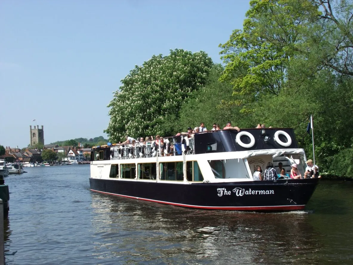 The Waterman boat has just departed the Hobbs of Henley boatyard. It is full of passengers enjoying one of the daily river trips. You can see Henley on Thames in the background, the trees are green and there are blue skies.