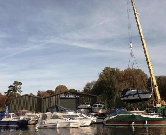 A boat is being lifted out of the river Thames in Henley to be stored at the Hobbs of Henley unit behind. There are a number of boats in the river, and it appears to be an autumn day.