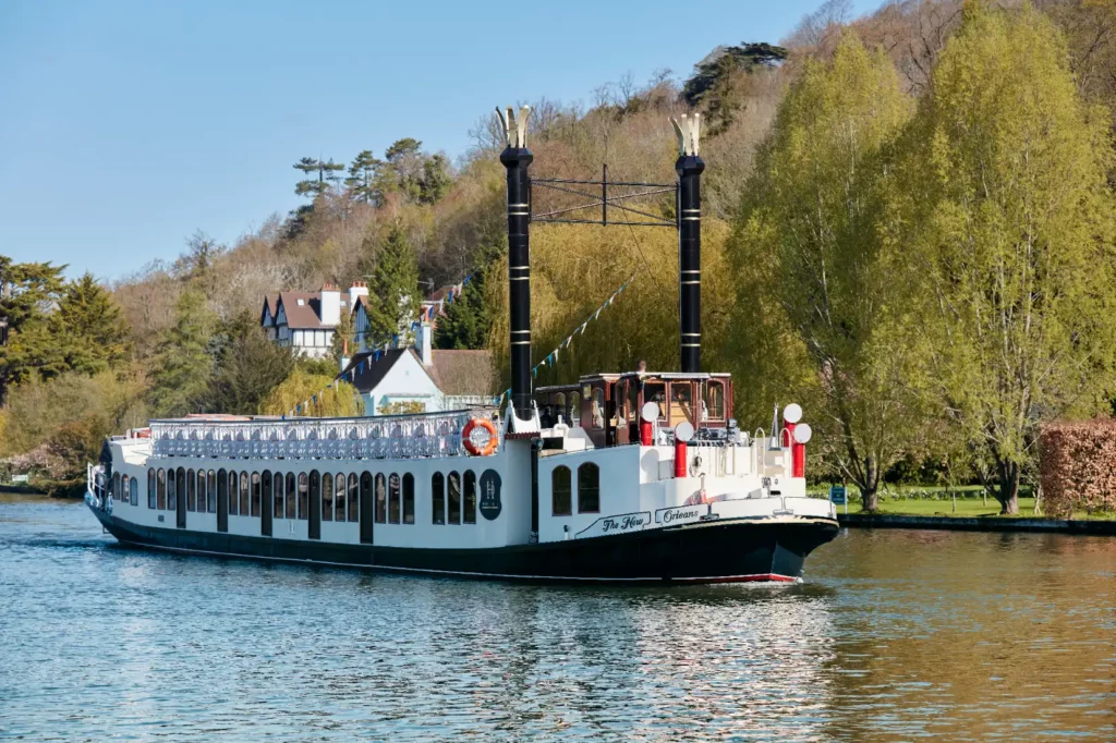 Hobbs of Henley boat The New Orleans cruising along the river thames, with a number of large houses in the background.