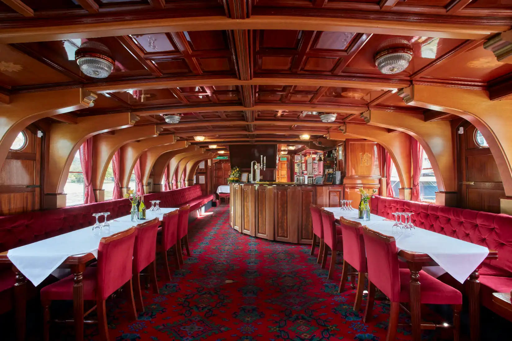 Inside the Hobbs of Henley boat The New Orleans, with wooden features, red seating and a red carpet. The tables are set up with white tablecloths.