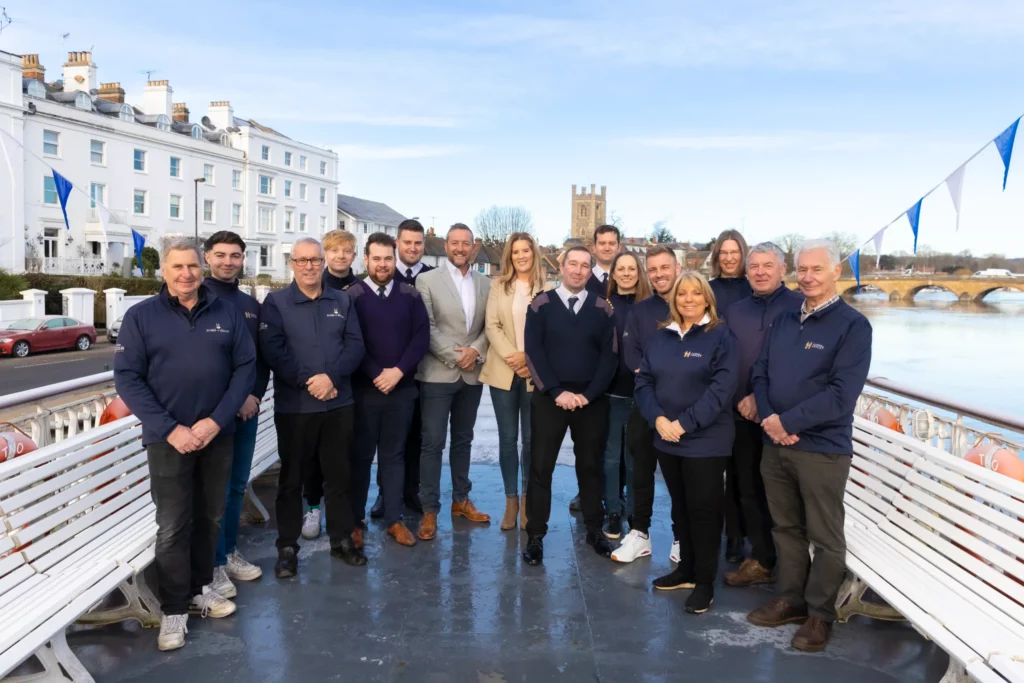 All of the Hobbs of Henley team stood on outside on one of their boats, with the river, bridge, and church in the background.