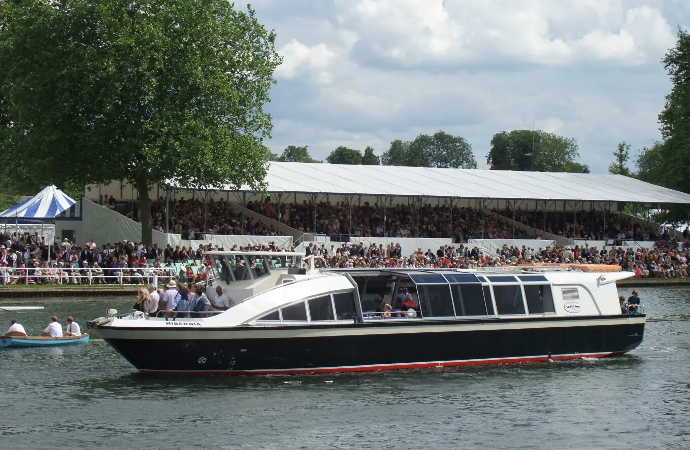 The Hibernia cruising along the river thames, with the Henley Royal Regatta busy crouds alongside the river and in the stands watching the rowing races.