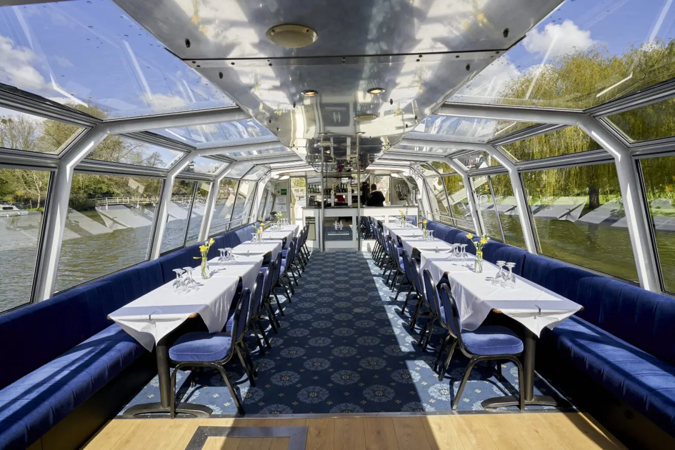 Inside the Hobbs of Henley boat Hibernia. The seating is blue velvet, and tables are laid with white tablecloths, glasses and yellow flowers. The sun is shining through the many glass windows.