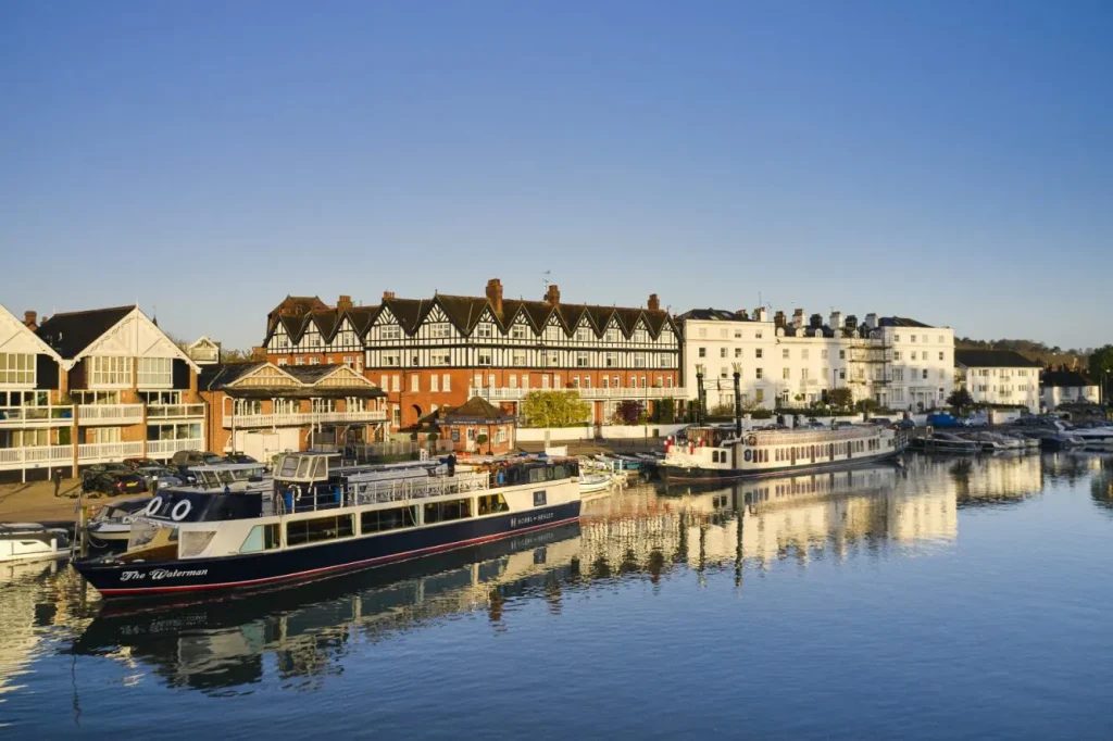 The Waterman and The New Orleans large passenger boats are pictured sat on the River Thames in Henley-On-Thames. Behind the boats is the Hobbs boatyard and nearby houses, the sky is blue and clear.