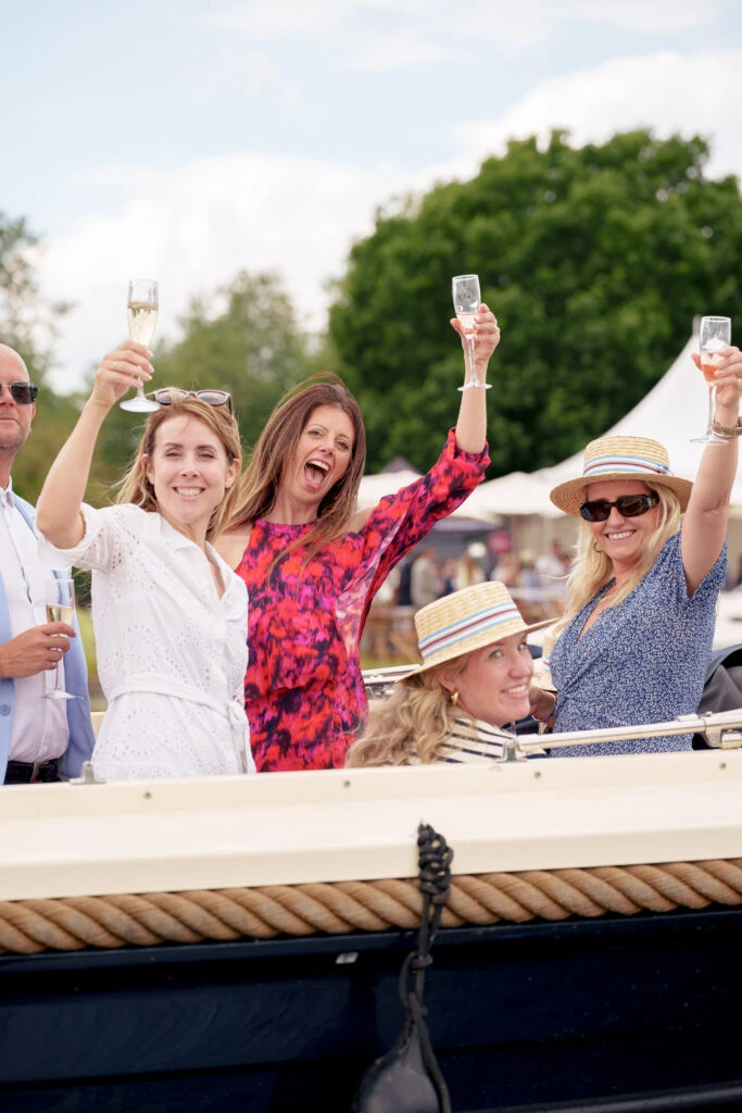 A group of 5 people aboard a Hobbs of Henley boat at the regatta. They are seen smiling, raising glasses of champagne, and having a wonderful time.